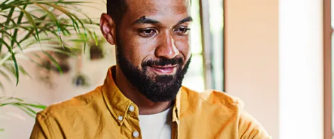 A man smiles while using a laptop in a sunny room filled with plants.