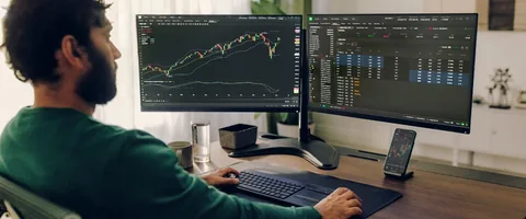 A trader viewing market charts and quotes on two monitors and a phone.
