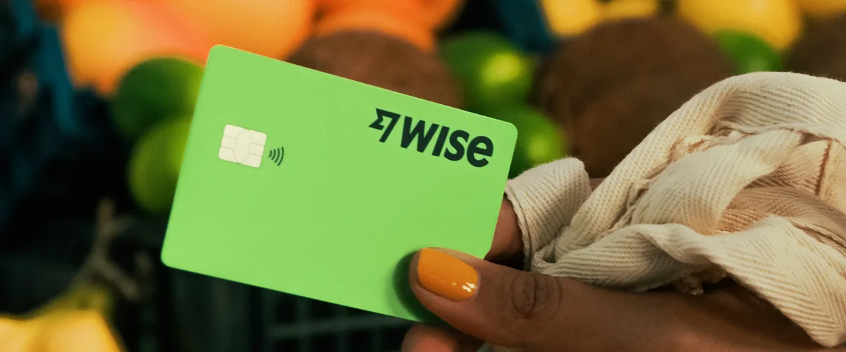 A hand holding a green Wise debit card in a produce aisle.